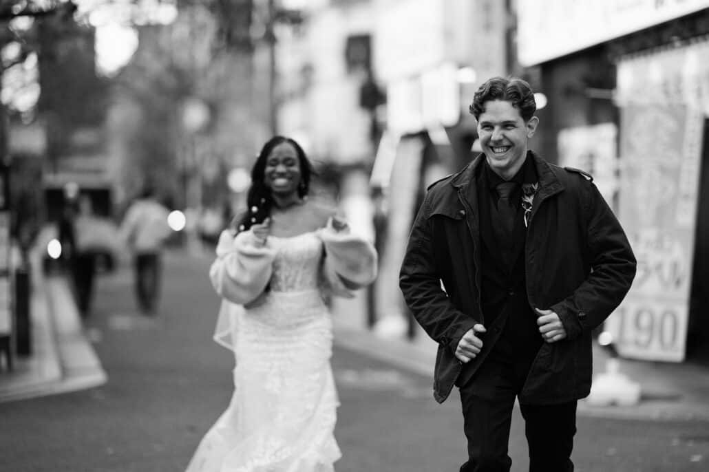 A black and white frame of the bride and groom laughing as they walk down a busy Kyoto street in their wedding clothes.