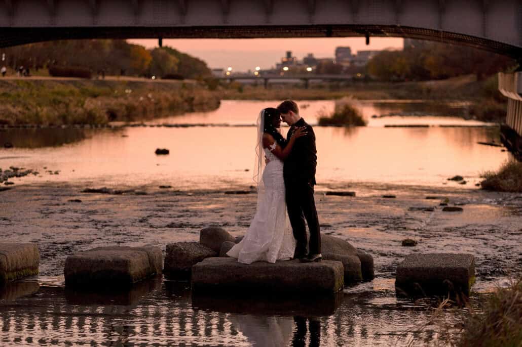 The couple embrace on the stepping stones of the Kamo River in Kyoto at sunset, a bridge overhead and a pink sky behind.