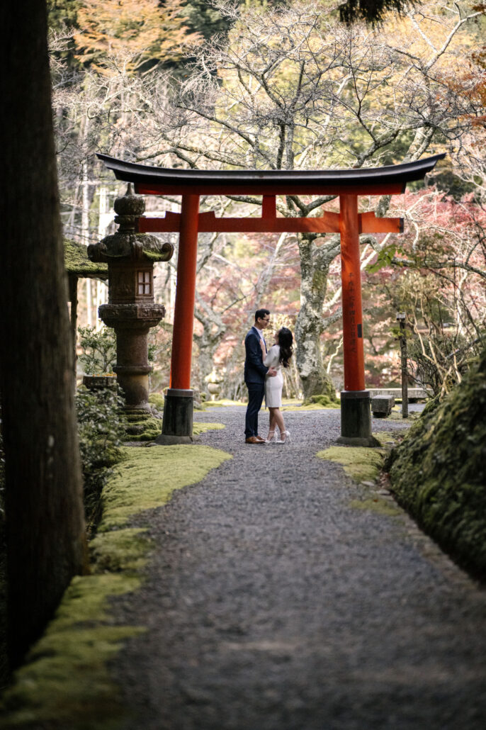 Framed by a cedar trunk and a mossy bank, the couple face each other beneath a red torii and stone lantern at their elopement in Kyoto.