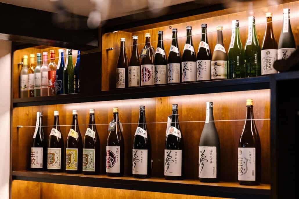 Rows of Niigata sake bottles with brush lettered labels, lit warmly on wooden shelves.