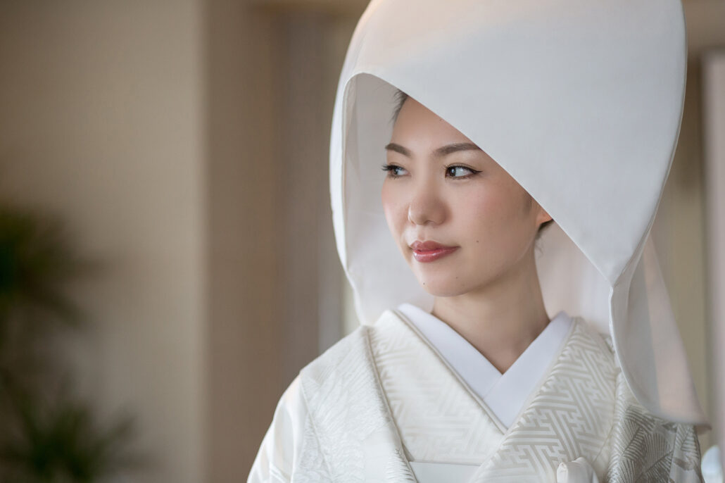A bride in a white wataboshi hood and patterned white shiromuku looks toward soft window light before her Shinto wedding in Tokyo