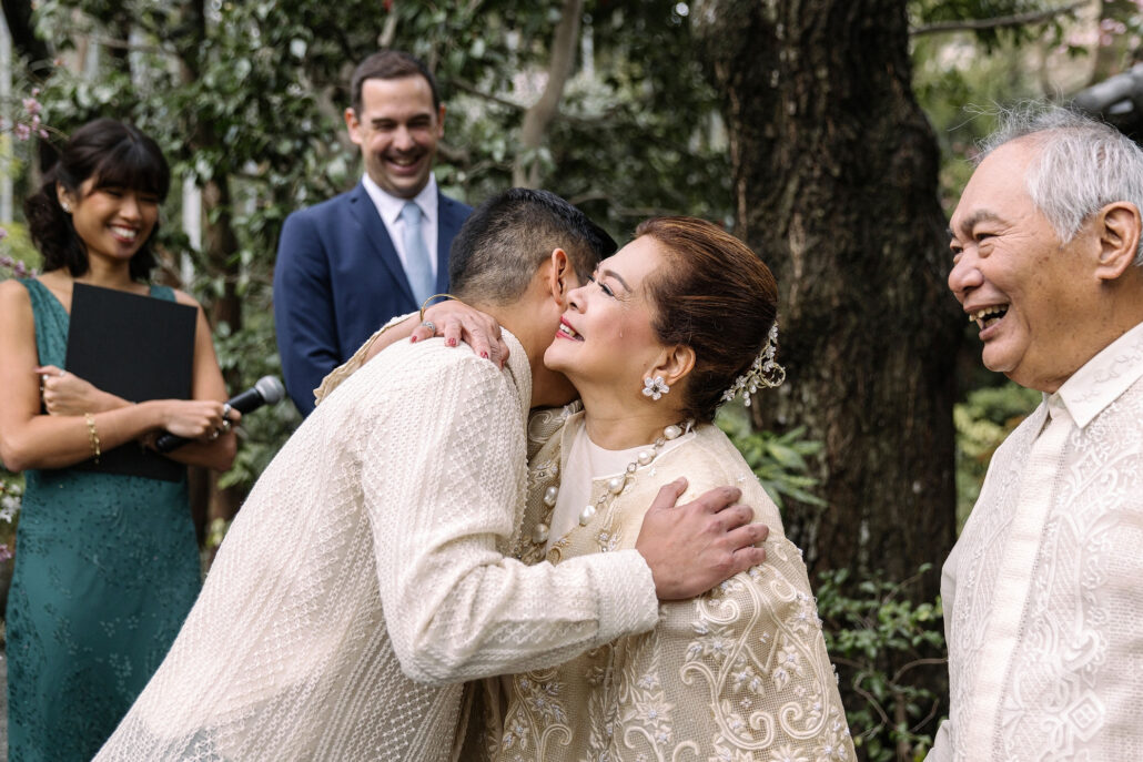 The groom embraces an older woman in an embroidered Filipiniana dress as family smile beside them at a wedding in Japan