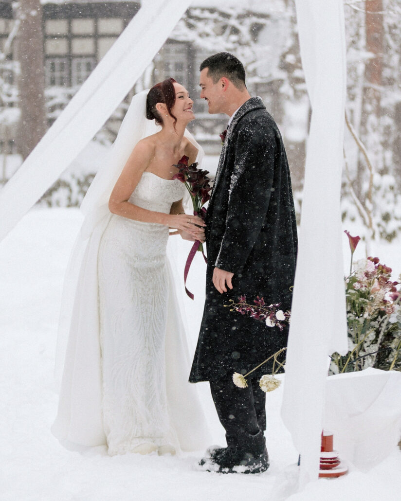 The bride and groom laugh together between white drapes at their forest ceremony in Hakuba, snow settling on his dark overcoat.