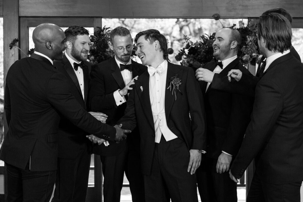 In black and white, the groomsmen crowd in to congratulate the groom, all laughing.