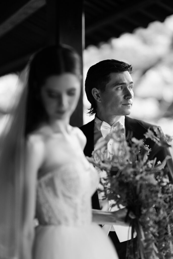 In black and white, the groom looks out at the garden while the bride looks down at her flowers.