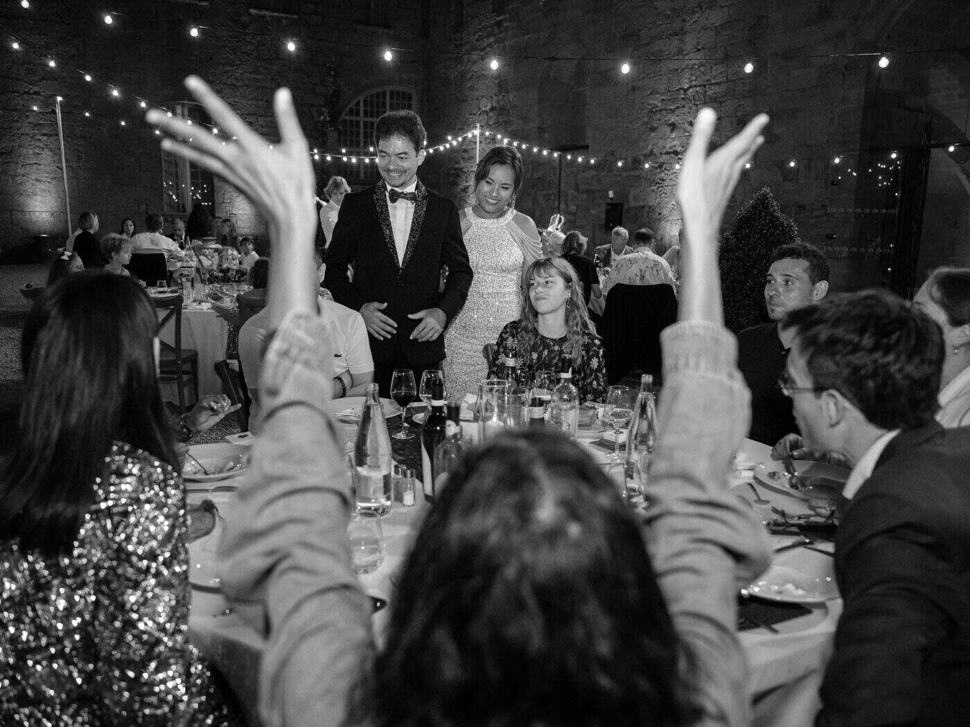 Black and white frame of guests raising their hands to cheer the newlyweds as they stand to speak at dinner in the abbey courtyard.