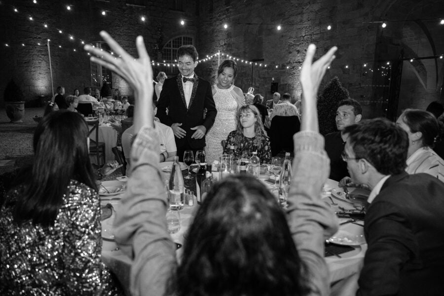 Black and white frame of guests raising their hands to cheer the newlyweds as they stand to speak at dinner in the abbey courtyard.