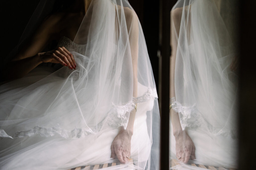 The bride's hand gathers her veil by the window, her reflection doubled in the glass.