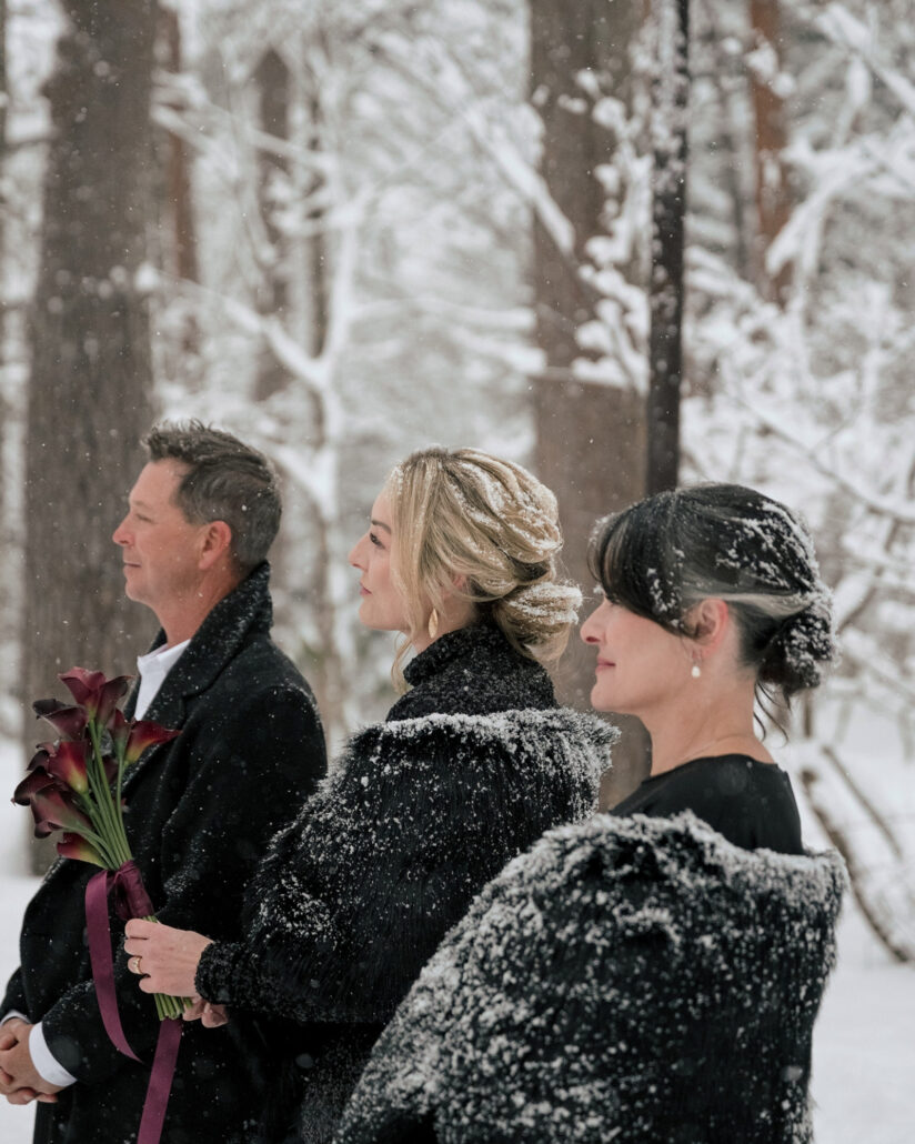 A man in a dark overcoat and two women in snow dusted fur wraps watch the vows, one holding the red calla lilies, at a snow wedding in the Hakuba forest.