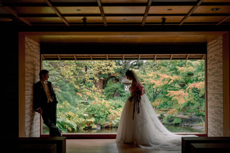 The couple framed by a wide opening onto the Japanese garden at Jinya, the groom leaning on the wall.
