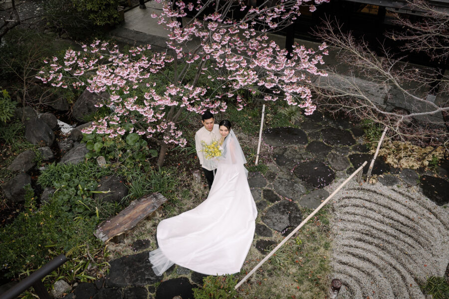The bride and groom stand beneath pink cherry blossom beside raked gravel in the garden at Motoyu Jinya, her train sweeping the stones.