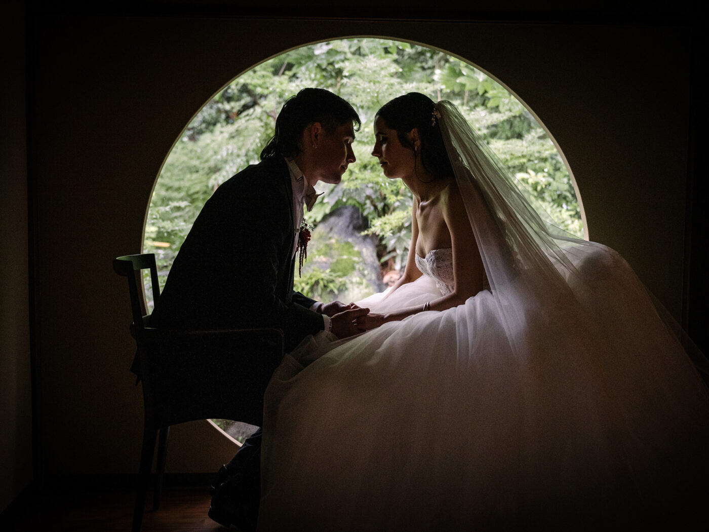 The bride and groom sit close in silhouette inside a round window looking out to the garden.