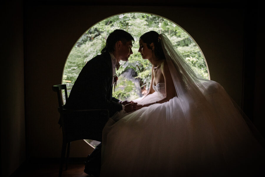 The bride and groom sit close in silhouette inside a round window looking out to the garden.