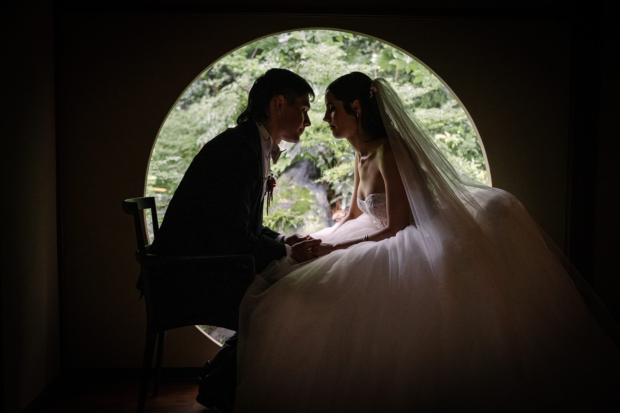 The bride and groom sit close in silhouette inside a round window looking out to the garden.
