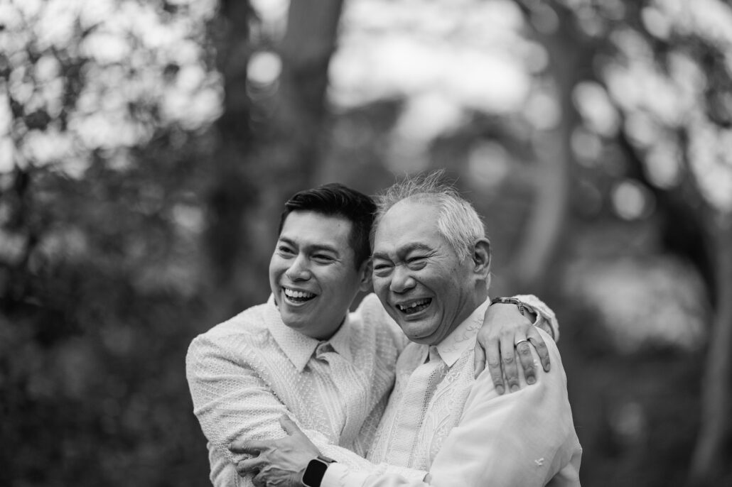 Black and white photograph of the groom laughing as an older relative hugs him close in the garden at his wedding in Japan