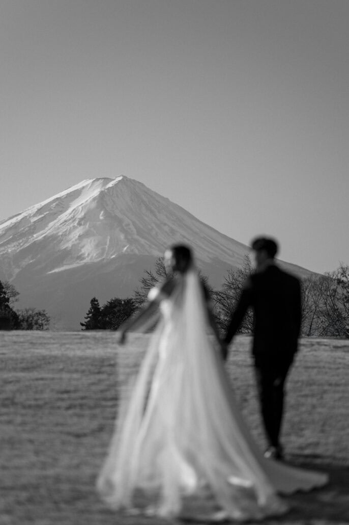A black and white frame of the bride's veil blowing out as she walks with the groom in front of Mt Fuji.