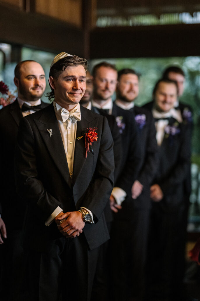 The groom stands at the front with his groomsmen, eyes on the bride.