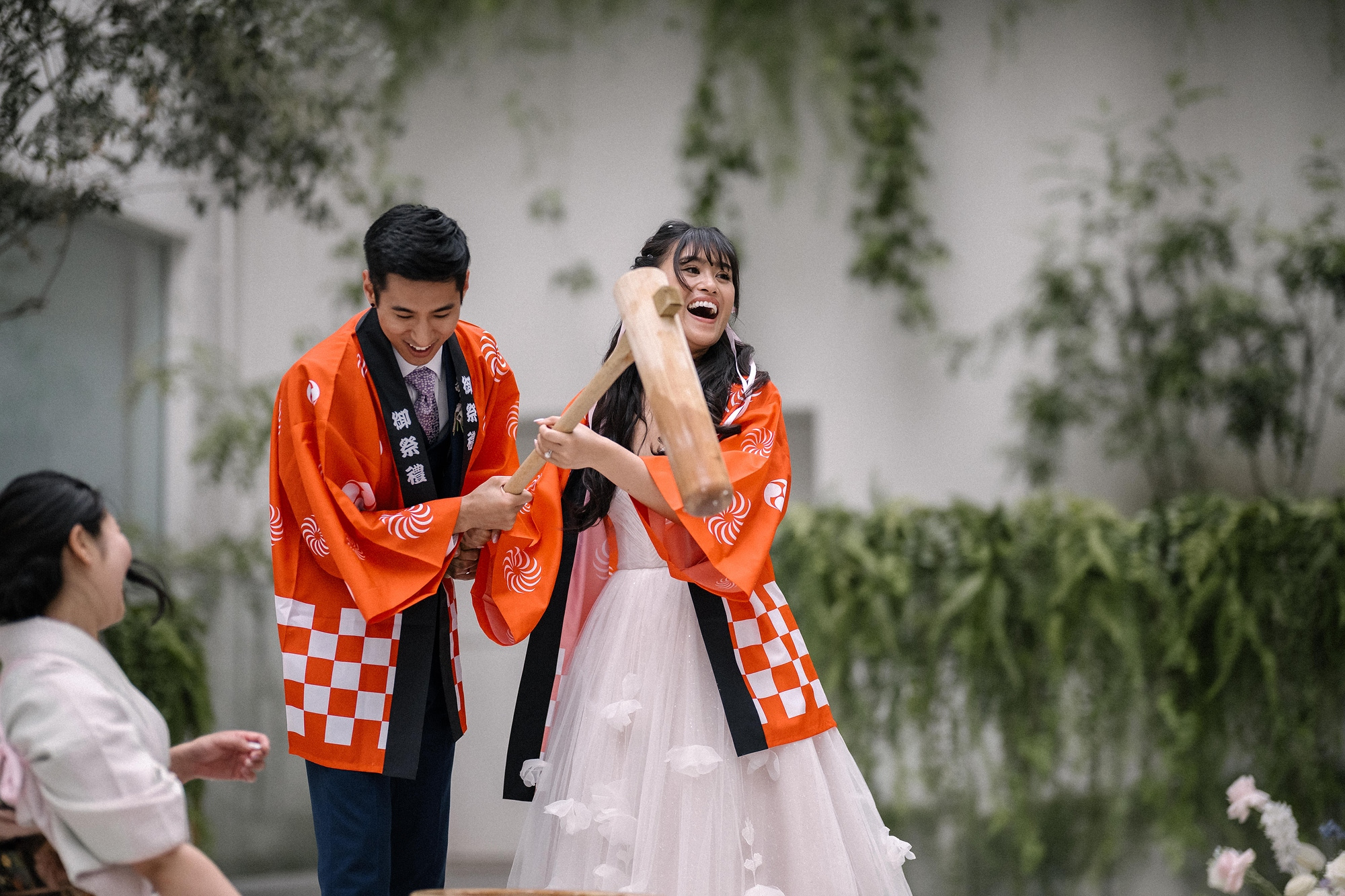 A bride and groom in orange happi coats hold a wooden mallet together over the mortar for mochi pounding at their wedding in Tokyo.