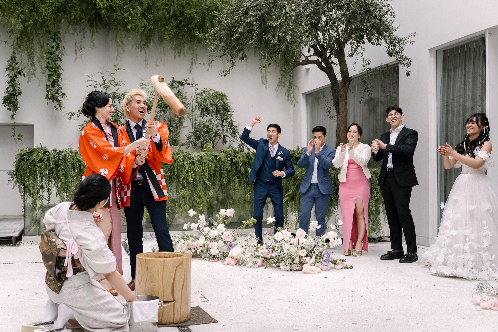 Guests in orange happi coats pound mochi with a wooden mallet in a white courtyard as the wedding party cheers, a Japanese wedding custom.