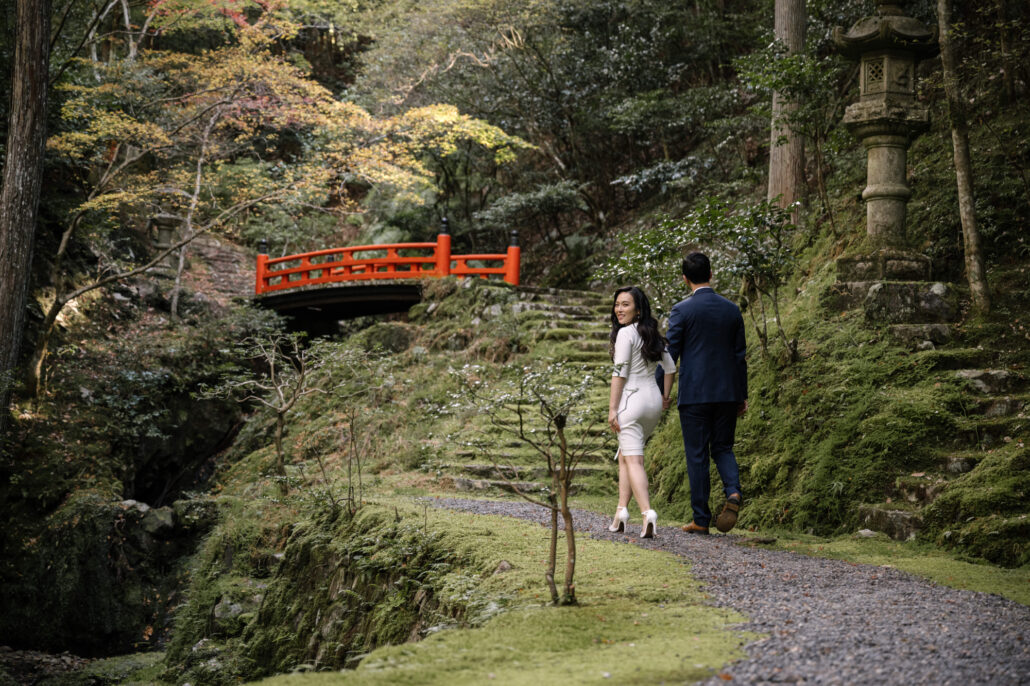 Walking hand in hand towards a vermilion bridge, she looks back and smiles, moss and a stone lantern on the slope beside them in Kyoto.