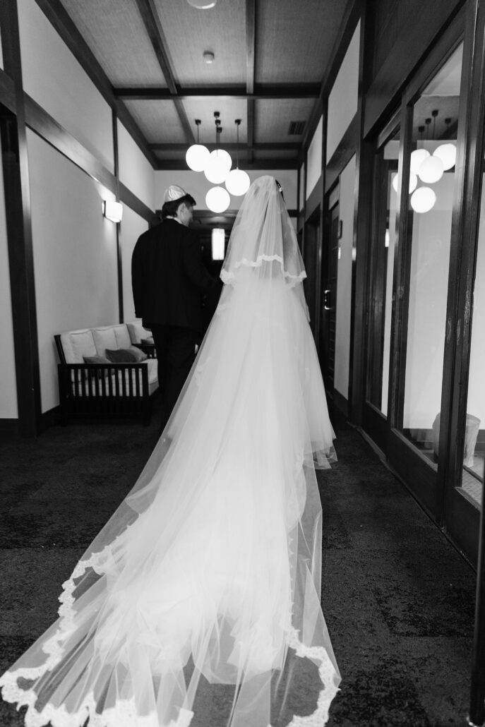In black and white, the bride's long train trails down a corridor behind the groom.