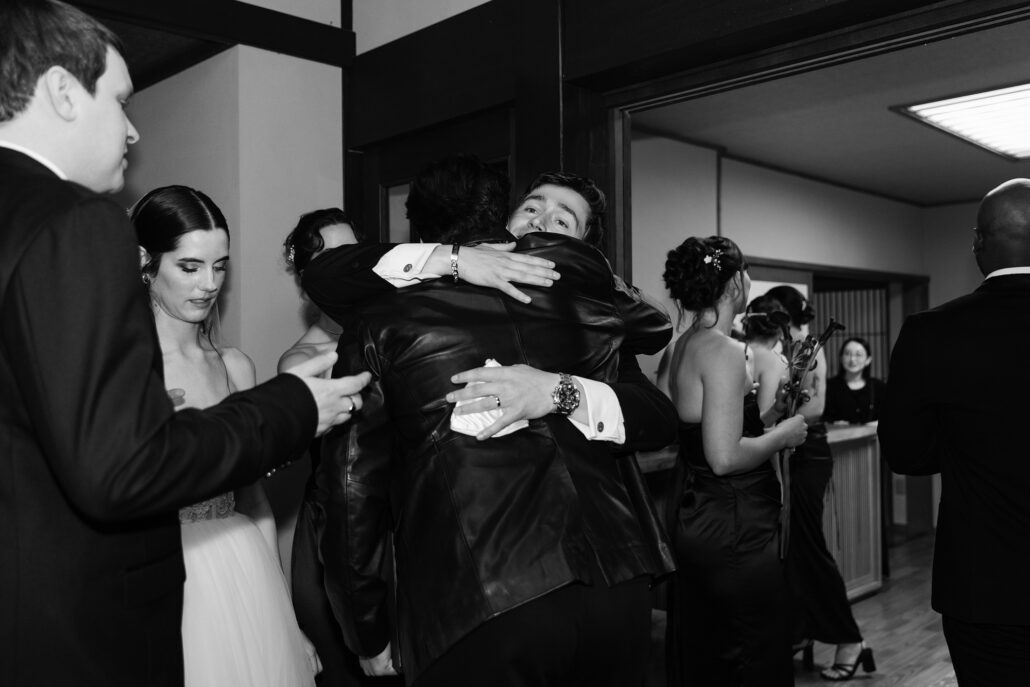 In black and white, a friend in a leather jacket hugs the groom as the bride stands beside them.