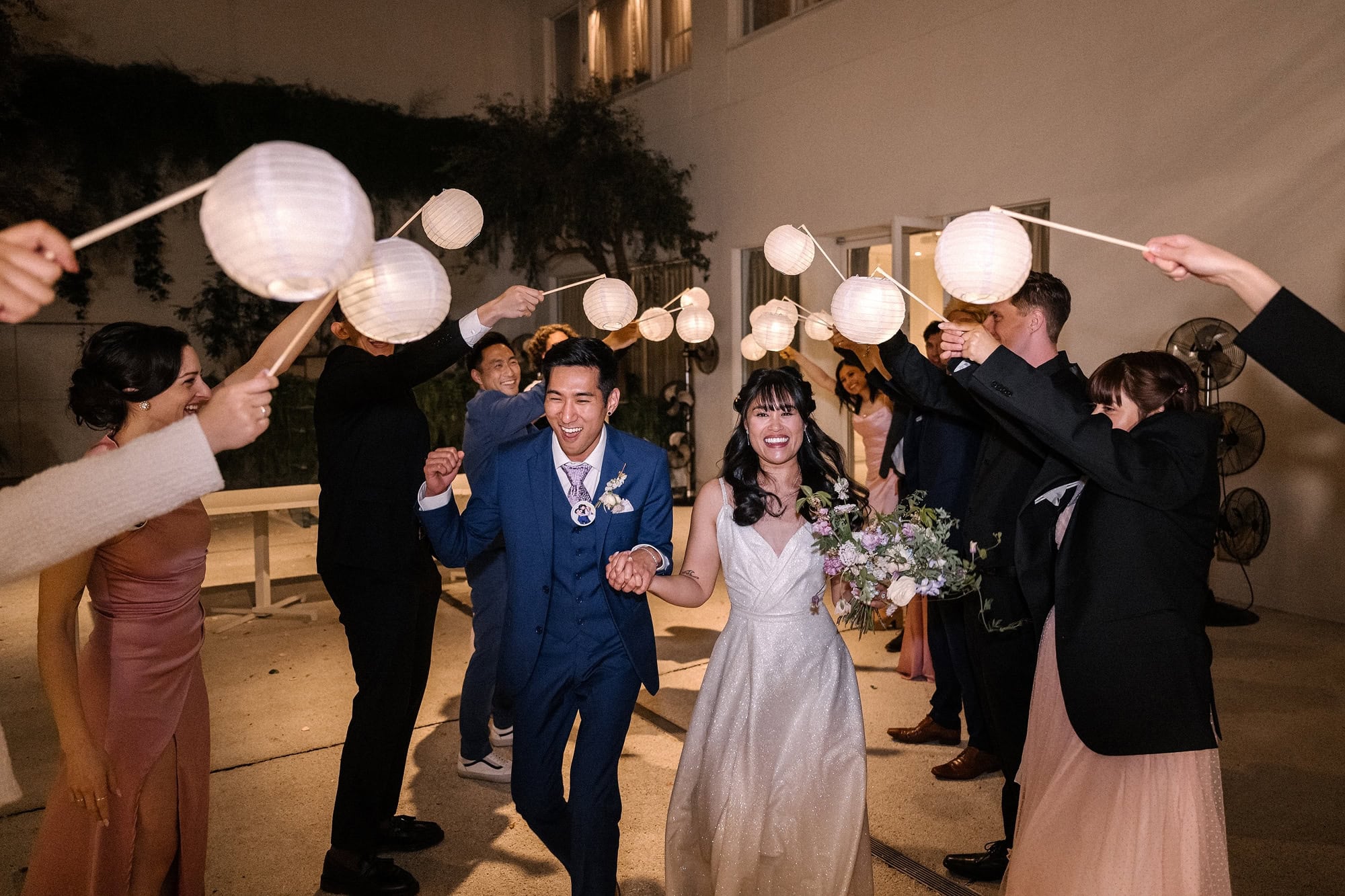 A bride and groom run laughing through a tunnel of white paper lanterns held high by their guests at night, the end of an intimate wedding in Tokyo.