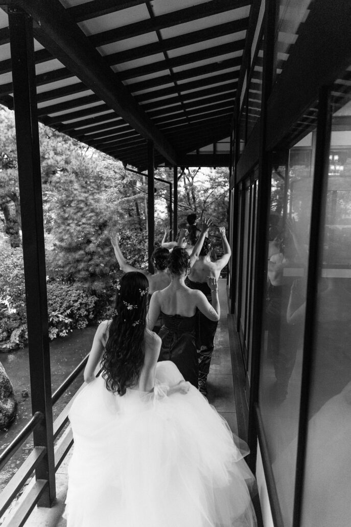 In black and white, the bride gathers her ball gown as she follows her bridesmaids down the covered veranda.