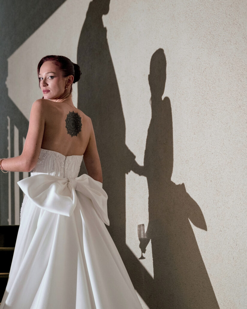 The bride looks back over her shoulder in a gown with a large bow as two guests cast shadows on the wall, one holding champagne, at a Japan destination wedding.