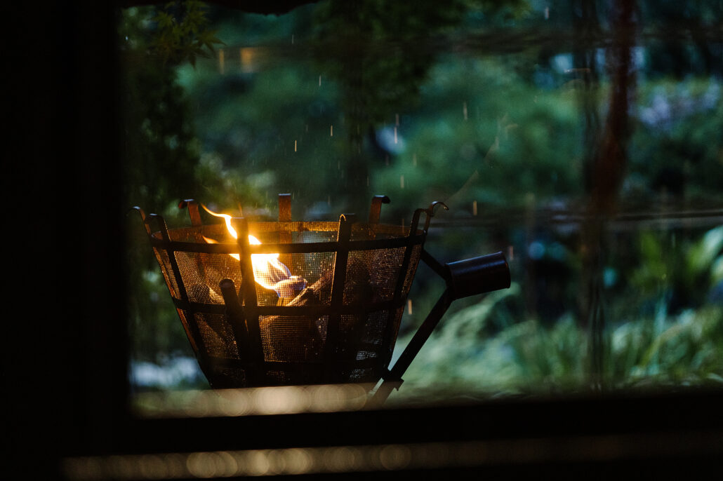 A candle burns in an iron lantern by a ryokan window at night, the Japanese garden dark beyond the glass