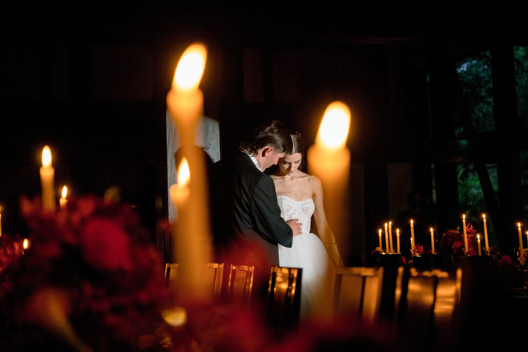The bride and groom behind tall candle flames, her gown catching the light.
