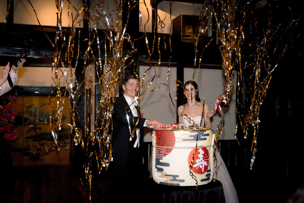 The couple at the kagami biraki barrel, mallets in hand, as gold and silver streamers fall in front of a gold folding screen.