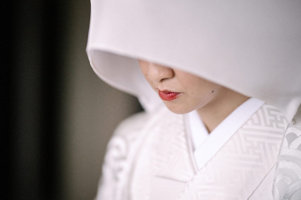 A close portrait of a bride beneath a white wataboshi hood, her red lips and white shiromuku collar lit by soft window light in Tokyo.