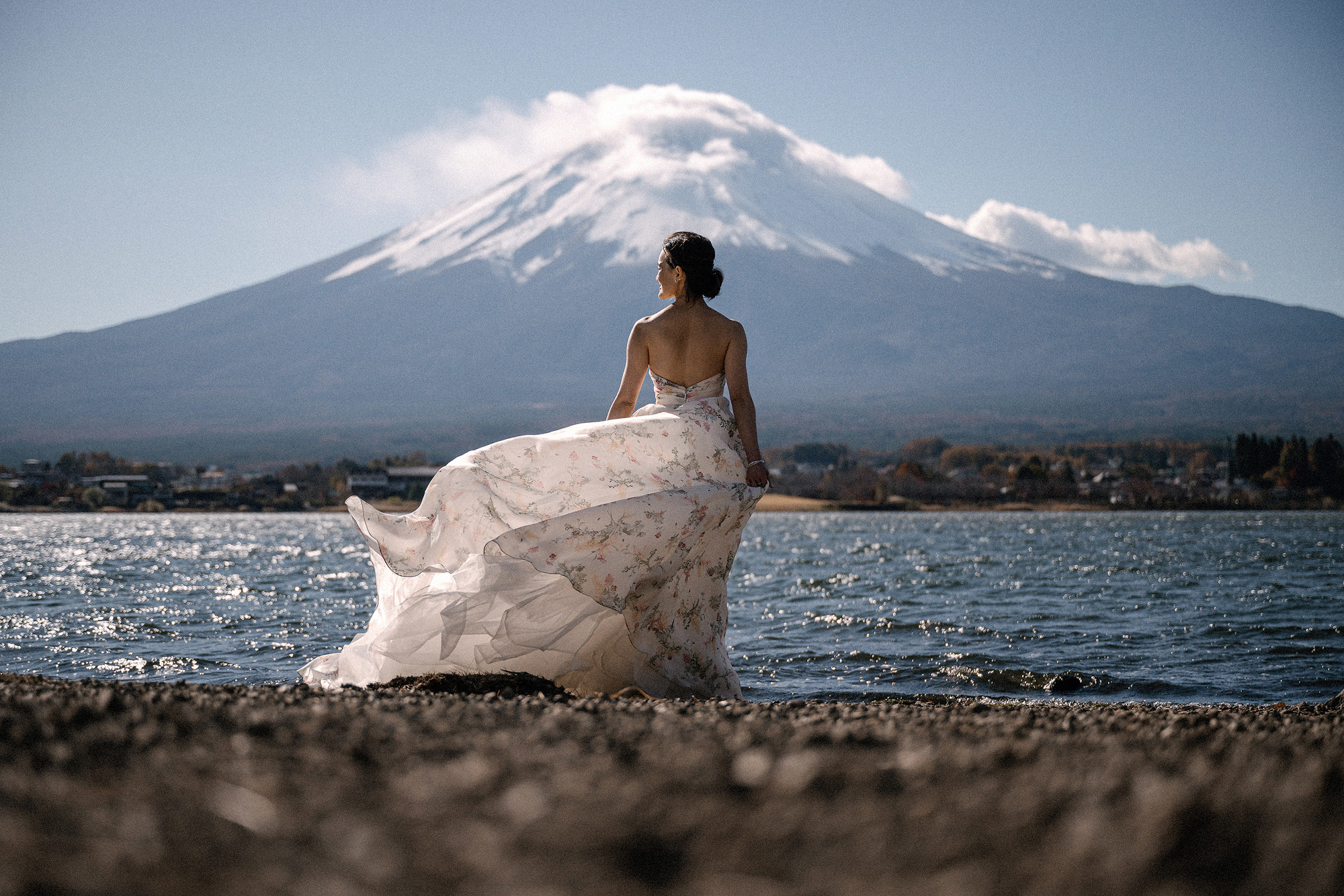 Her back to the camera, the bride's floral gown blows out in the wind on the shore of Lake Kawaguchi, Mt Fuji wearing a cap cloud behind her.
