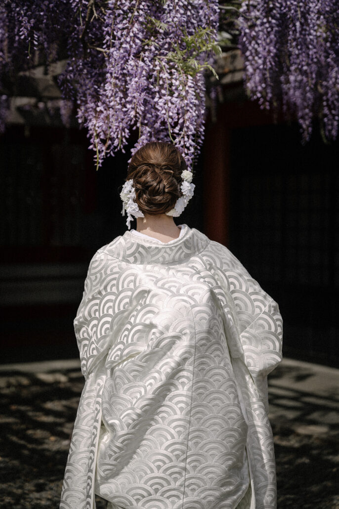 Seen from behind, a bride in a white shiromuku kimono patterned with waves, her hair dressed with flowers, stands beneath hanging purple wisteria.