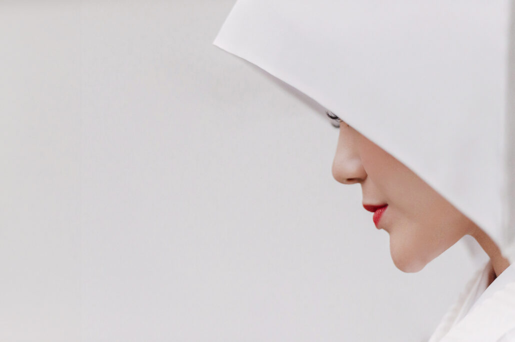 In profile, a bride's face and red lips frame the curve of a white wataboshi hood against a pale background, a quiet bridal portrait.
