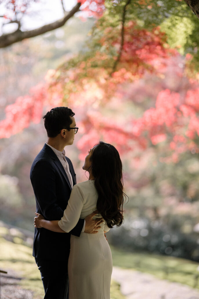 The couple hold each other beneath a maple branch, red leaves bright behind them, in a private garden in Kyoto in autumn.