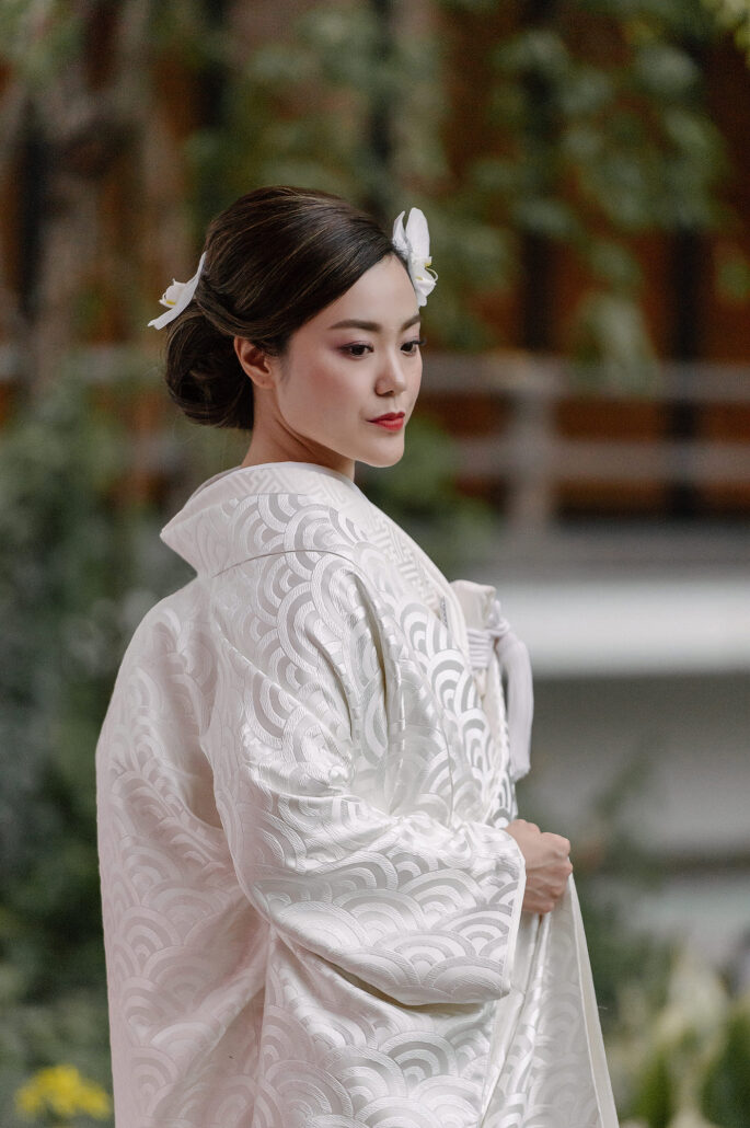 A bride in a white kimono patterned with seigaiha waves and a flower in her hair stands in a garden for her pre-wedding photos in Japan.