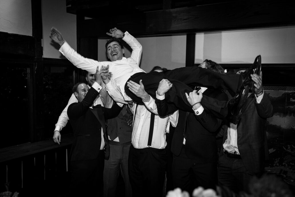 In black and white, the groom is hoisted above his friends, laughing.