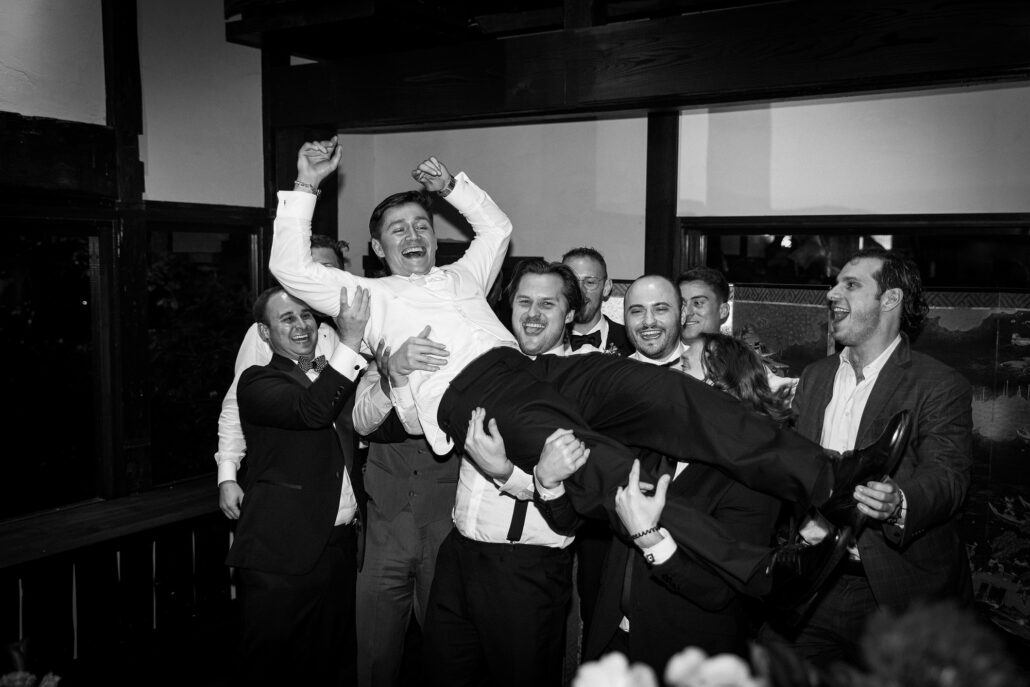 In black and white, friends hold the groom aloft as the room cheers.
