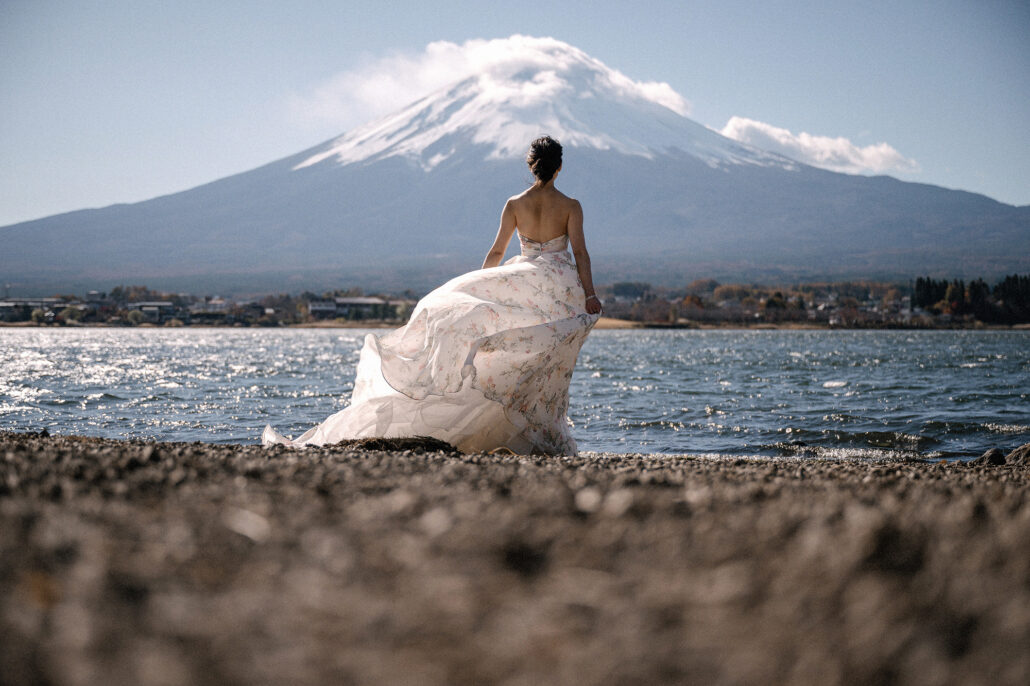 The bride's floral gown fans out in the wind on the stony shore of Lake Kawaguchi, Mt Fuji beyond the water.