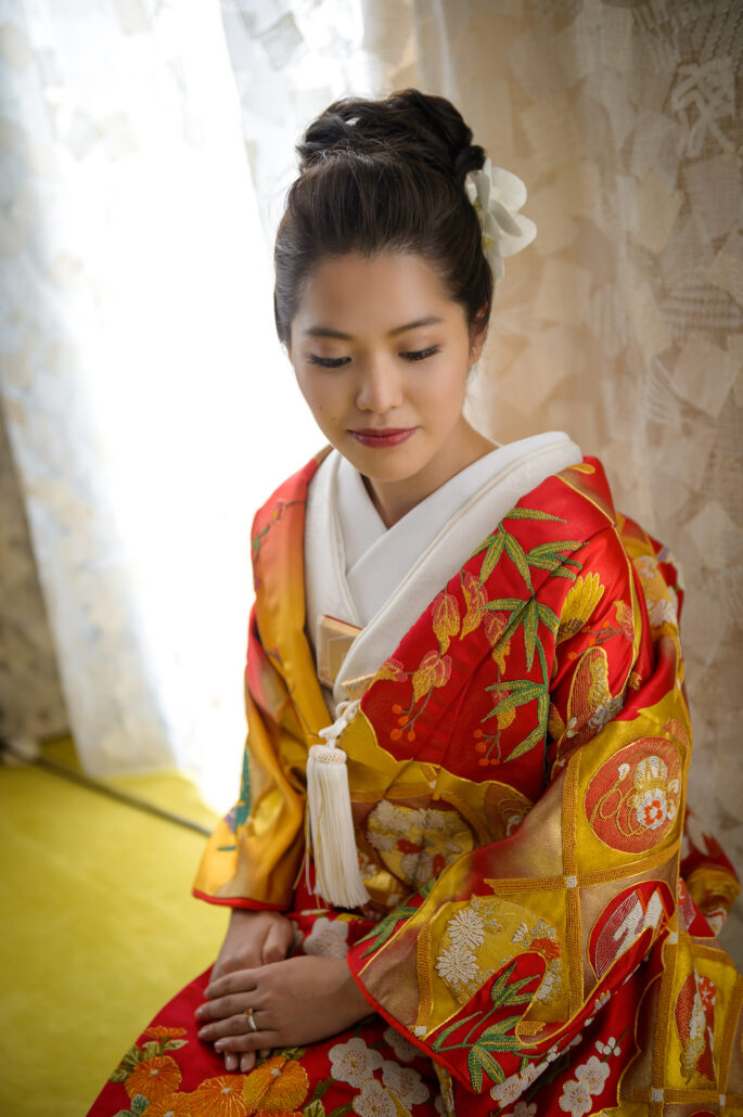 A bride in a red and gold iro-uchikake embroidered with bamboo, blossoms and fans sits in soft window light, a white flower in her hair.
