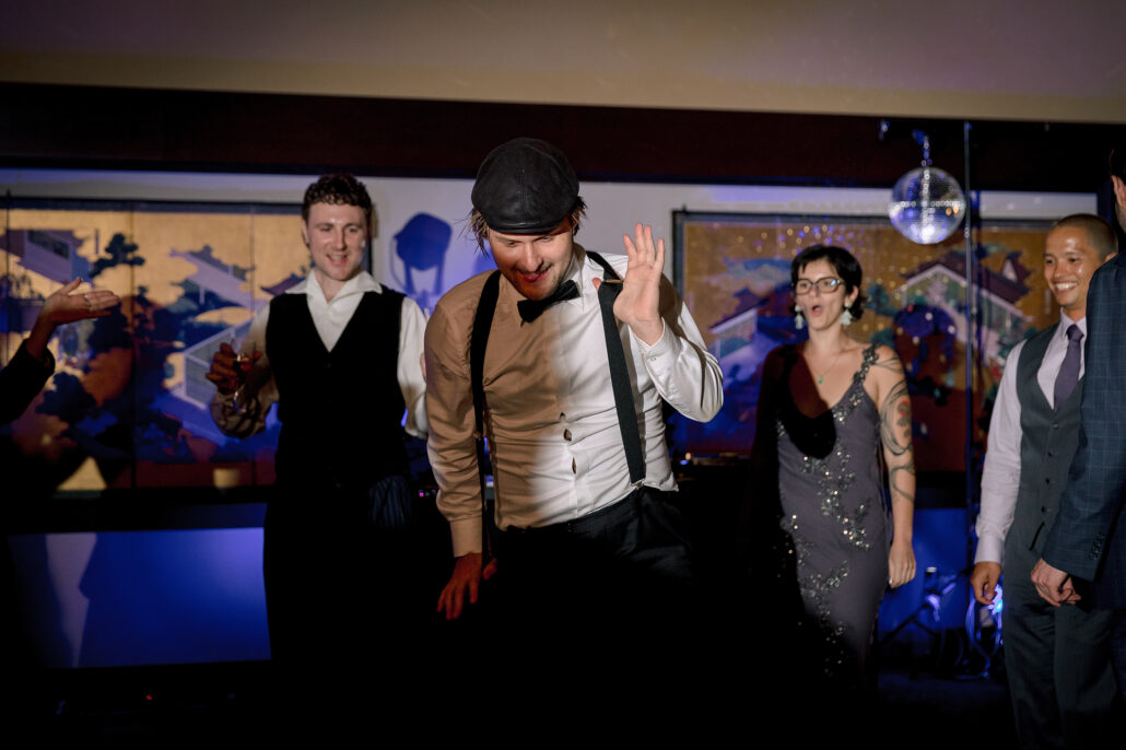 Guests cheer from the edge of the dance floor, one in braces and a flat cap.