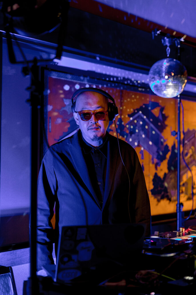 The DJ in sunglasses at the decks under blue light and a mirror ball.