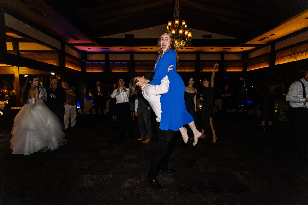 A guest in a blue dress is lifted mid-dance as the bride dances nearby.