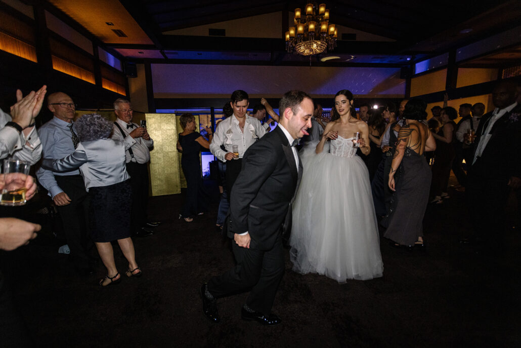 A guest in a dark suit dances low across the floor toward the bride as guests clap along.