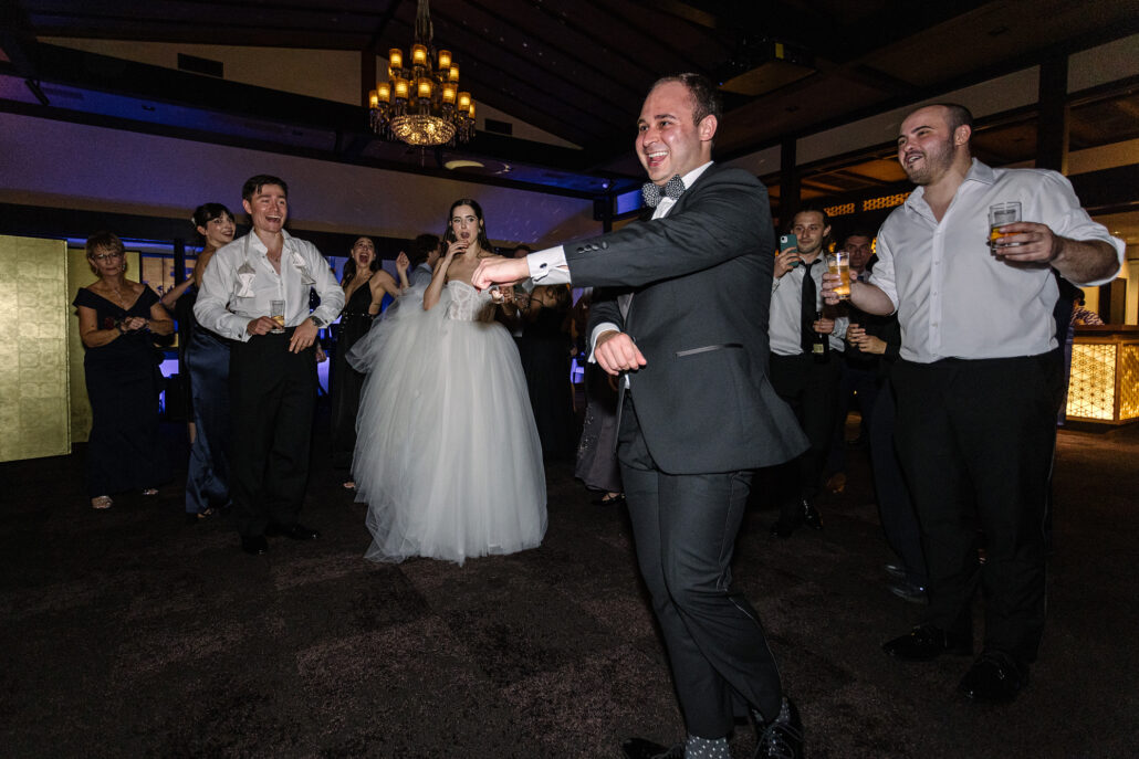 A guest in a grey suit dances out in front as the bride watches behind him.