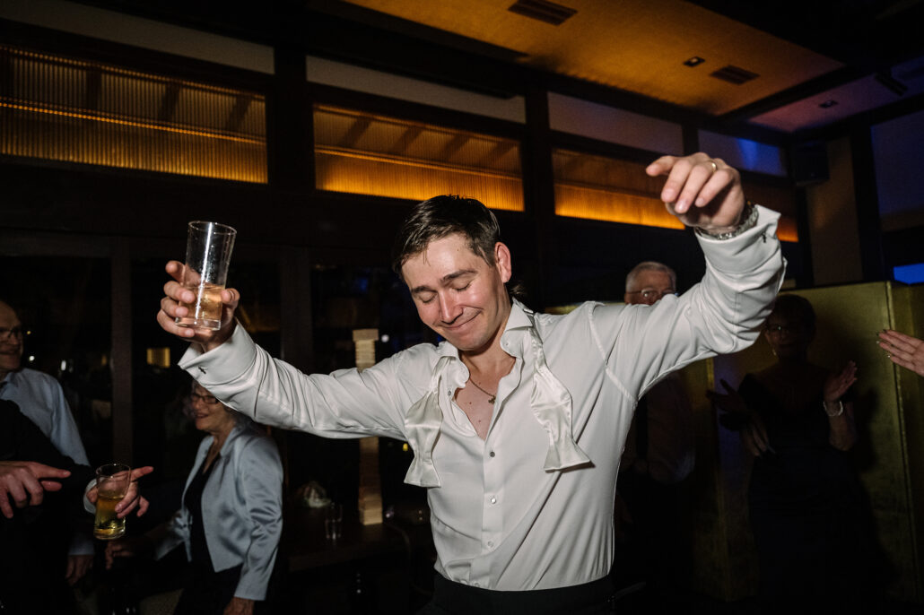 The groom dances with his glass raised and his collar open.