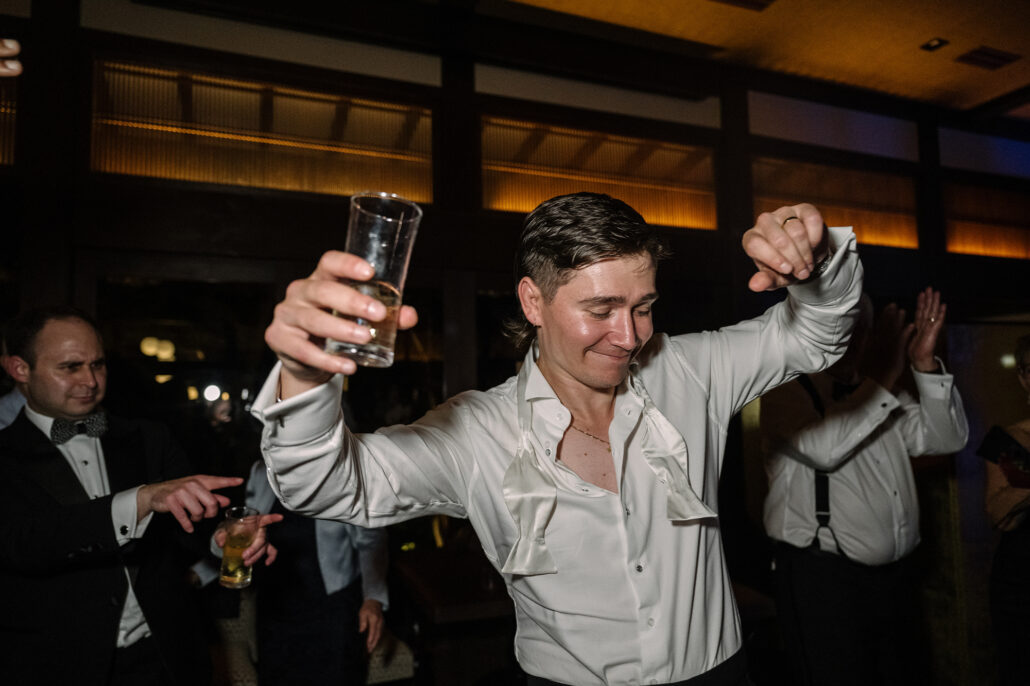The groom dances with both arms up, a glass in one hand, guests behind.
