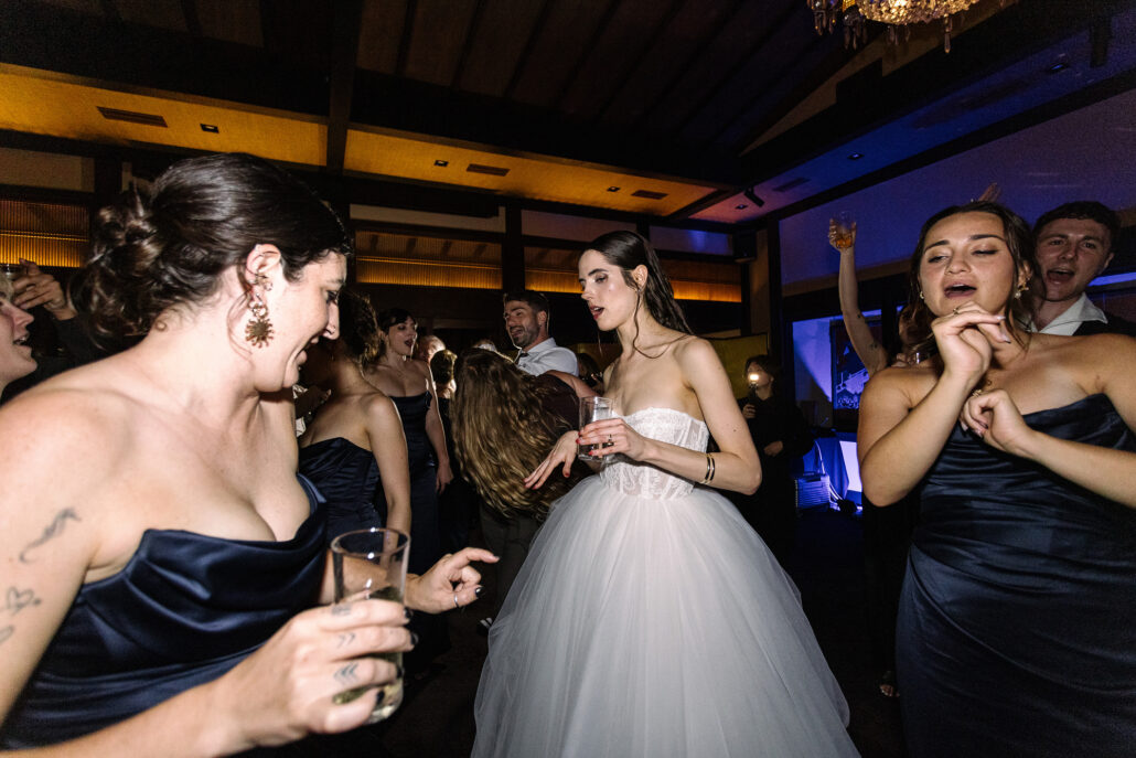 The bride dances among her bridesmaids, glasses in hand.